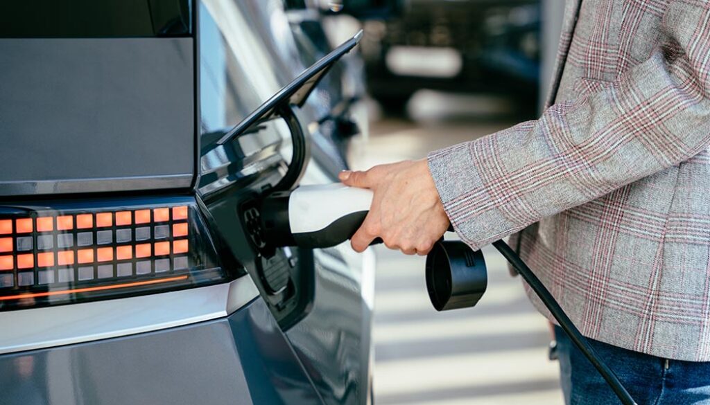 Woman holding charging cable for electric car , looking happ waiting electric car to charge. Caucasian female stands near electric auto in dealership. Smart ecological living