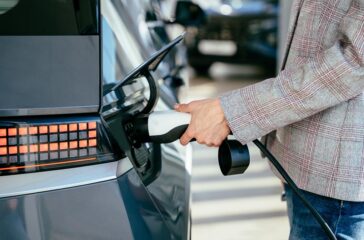 Woman holding charging cable for electric car , looking happy waiting electric car to charge. Caucasian female stands near electric auto in dealership. Smart ecological living