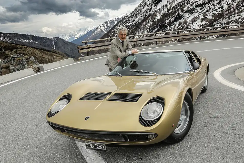 Stardesigner Marcello Gandini lehnt an einem goldenen Lamborghini Miura vor einer alpinen Kulisse – Ein Meisterwerk des Bertone-Designs.