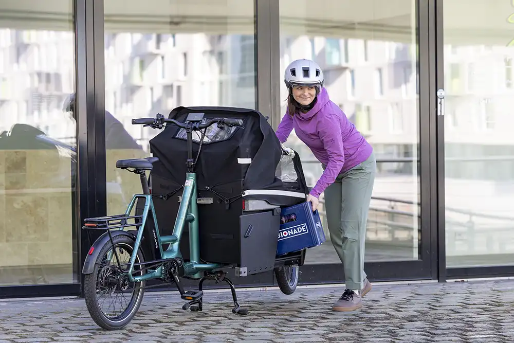Praktischer Alltagshelfer: Eine Frau belädt das i:SY Cargo Maxi mit Getränkekisten; sicherer Stand durch Zweibeinständer.