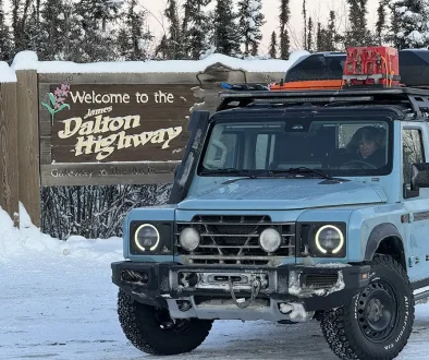 Ein babyblauer Ineos Grenadier mit Dachträger und Expeditionsausrüstung vor dem hölzernen Willkommensschild des James Dalton Highway in einer verschneiten Winterlandschaft in Alaska.