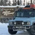 Ein babyblauer Ineos Grenadier mit Dachträger und Expeditionsausrüstung vor dem hölzernen Willkommensschild des James Dalton Highway in einer verschneiten Winterlandschaft in Alaska.