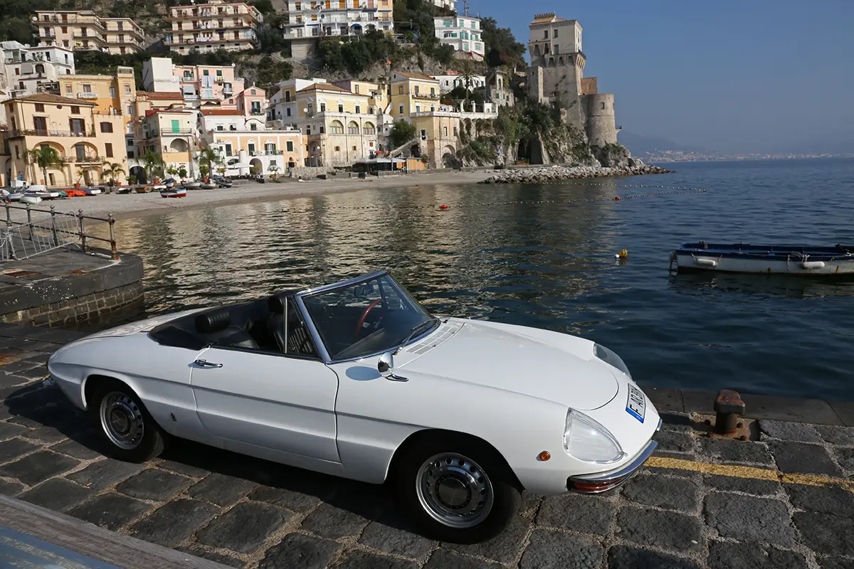 Weisser Alfa Romeo Spider Duetto parkiert am Hafen vor einer malerischen italienischen Stadtkulisse.