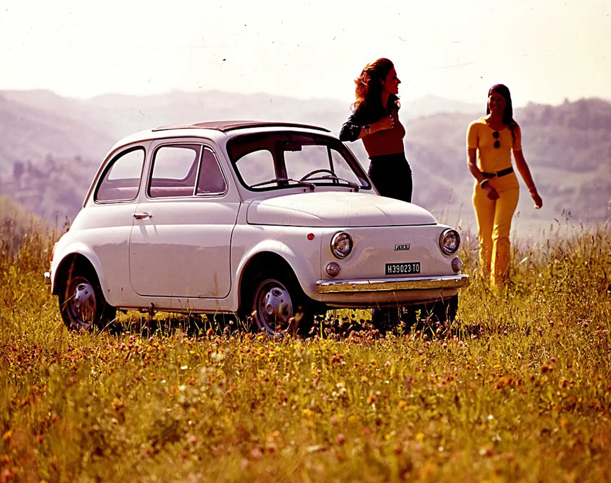 Ein klassischer weißer Fiat Nuova 500 mit geöffnetem Faltdach auf einer Wiese in Italien, im Hintergrund zwei Personen.