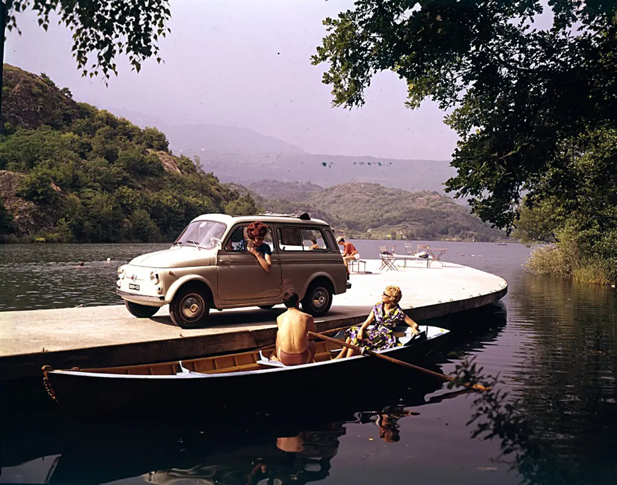 Ein hellbeiger Fiat Nuova 500 Giardiniera Kombi des Modelljahrs 1960, geparkt auf einem Steg direkt an einem idyllischen italienischen See mit einem Boot im Vordergrund.