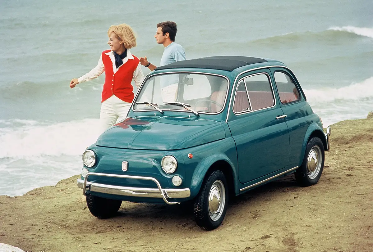 Ein dunkelgrüner Fiat 500 DeLuxe aus dem Jahr 1968, abgestellt auf sandigem Untergrund direkt am Meeresstrand mit einem Paar im Hintergrund.