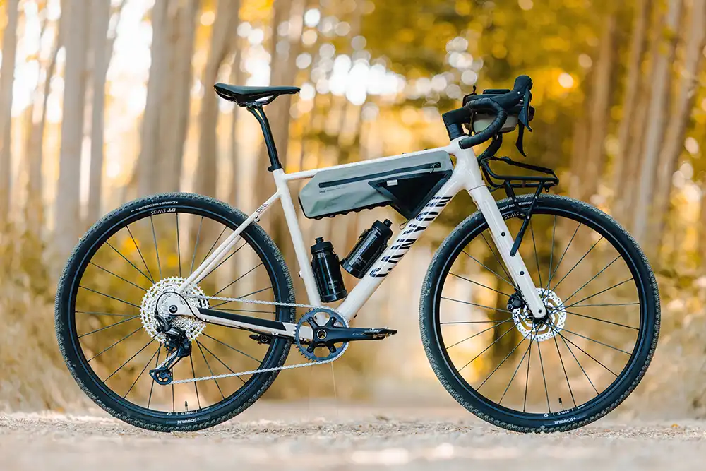 Das neue Canyon Grizl AL Gravel-Bike in Silber-Grau mit montierter Rahmentasche, zwei Trinkflaschen und einem Frontgepäckträger, perfekt in Szene gesetzt in einem herbstlichen Waldgebiet.
