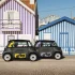 Zwei schwarze Citroën Ami Rip Curl Sondermodelle mit gelben und lilafarbenen Design-Applikationen vor farbenfrohen, gestreiften Strandhäusern.