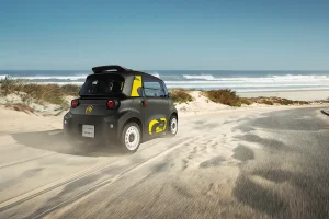 Small black compact car with yellow graphics driving along a sandy beach road with the ocean in the background.