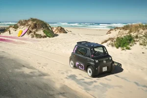 Small black compact car with pink decals driving on a sandy beach toward the ocean, surfboards leaning on a dune nearby.
