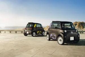 Two compact electric cars with colorful decals parked on a paved seawall, with the beach and dunes in the background under a clear sky.