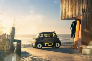 Compact black car with yellow graphics parked on a sandy beach beside a wooden beach shack with surfboards nearby at sunset.