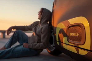 Person sitting on the ground at sunset, leaning against a graphic-painted car with Rip Curl branding on the door panel.