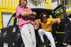 Two friends pose beside a black, yellow-accented car in front of a bright yellow striped building.