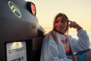 Woman sits beside a dark car at sunset, hair blown by the breeze, wearing a light sweatshirt and shorts by the beach. License plate shows Citroën x Rip Curl collaboration.
