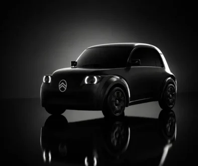 Ein im Dunkeln gehaltenes Teaser-Bild der Silhouette des kommenden elektrischen Citroën 2CV, das die kompakte Form des Kleinstwagens sowie markant leuchtende Frontscheinwerfer und das illuminierte Markenlogo zeigt.