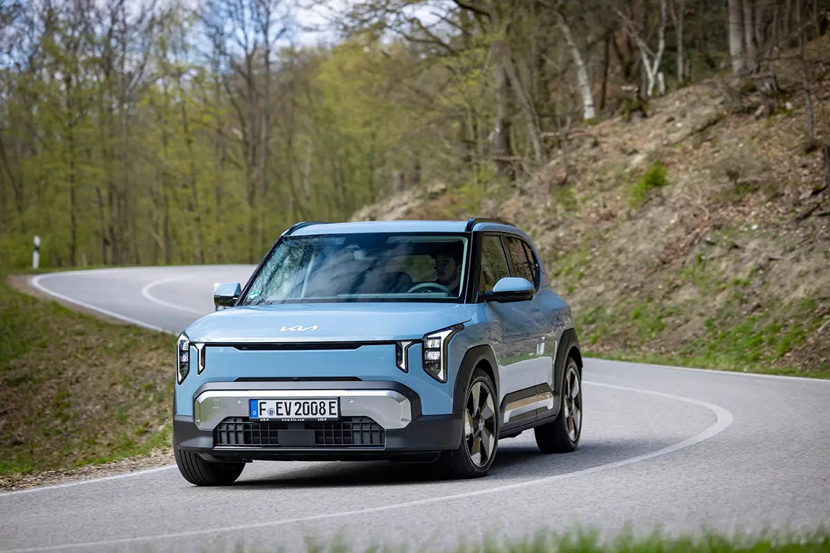 Hellblauer Kia EV2 im Crossover-Design auf einer kurvigen Straße vor herbstlicher Waldkulisse.
