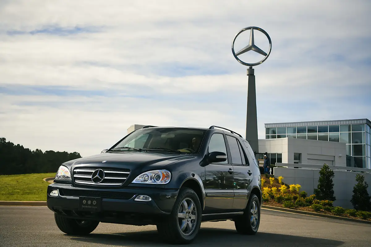 Ein klassischer, dunkler Mercedes-Benz ML vor dem Hauptgebäude von Mercedes-Benz US International in Tuscaloosa mit dem großen, hoch aufragenden Mercedes-Stern im Hintergrund.