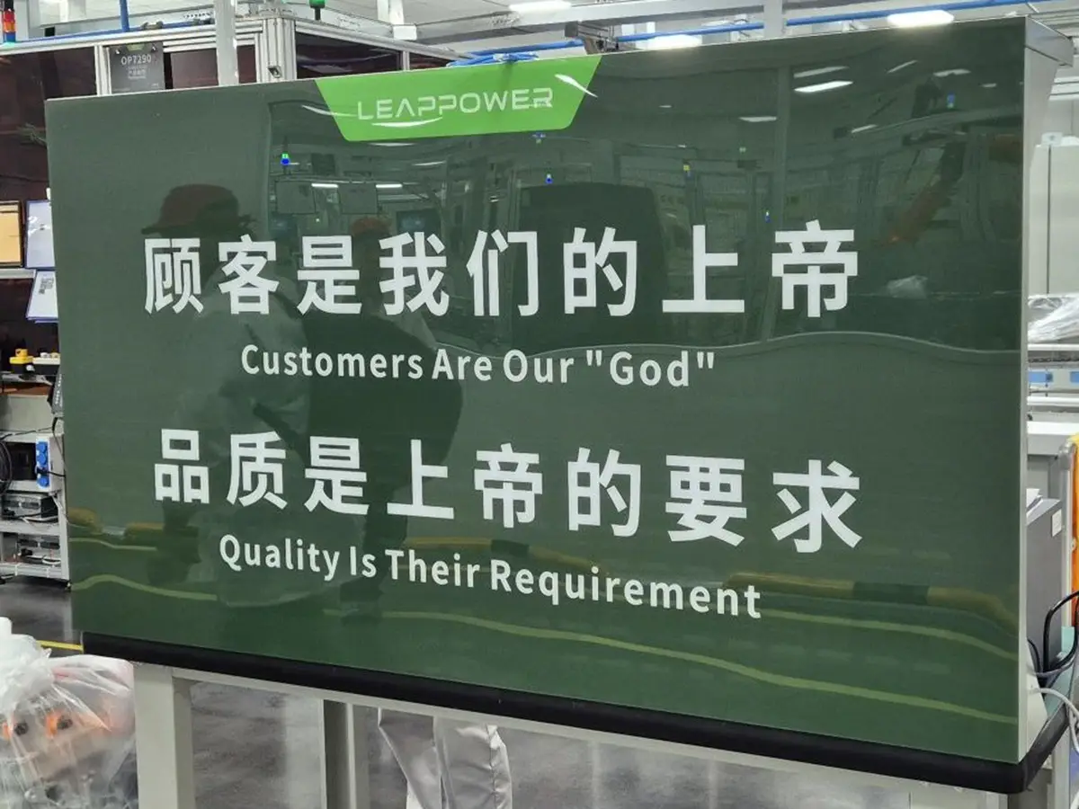 Ein grünes Schild in der Produktionsstätte mit Motivations-Sinnsprüchen auf Chinesisch und Englisch wie „Customers Are Our 'God' / Quality is Their Requirement“.