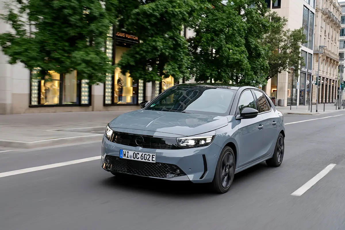 Grauer Opel Corsa Electric fährt auf einer städtischen Straße an Allee-Bäumen vorbei.