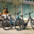 Zwei Personen in einer urbanen Umgebung sitzend, im Vordergrund stehen zwei Tenways Ago Performance Pedelecs mit aufgeräumter Optik.