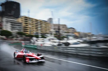 Red open-wheel race car speeding on a wet city harbor street, with blurred buildings and moored yachts in the background, spray behind the tires.