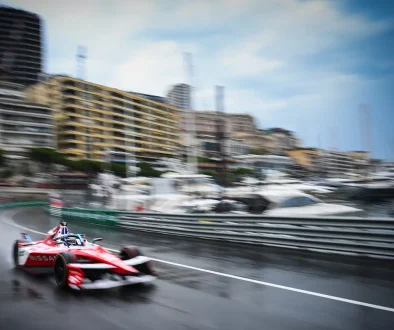 Red open-wheel race car speeding on a wet city harbor street, with blurred buildings and moored yachts in the background, spray behind the tires.