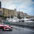 Red open-wheel race car speeding on a wet city harbor street, with blurred buildings and moored yachts in the background, spray behind the tires.
