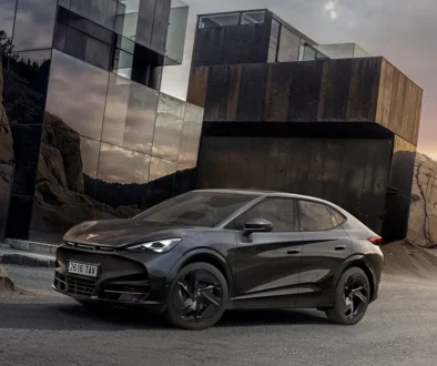 Der rein elektrische Cupra Tavascan Black Edition in der tiefschwarzen Lackierung Midnight Black und mit schwarzen 21-Zoll-Alufelgen parkt auf einer asphaltierten Strasse vor einer modernen, kantigen Gebäudearchitektur aus dunklem Metall.