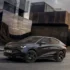 Der rein elektrische Cupra Tavascan Black Edition in der tiefschwarzen Lackierung Midnight Black und mit schwarzen 21-Zoll-Alufelgen parkt auf einer asphaltierten Strasse vor einer modernen, kantigen Gebäudearchitektur aus dunklem Metall.