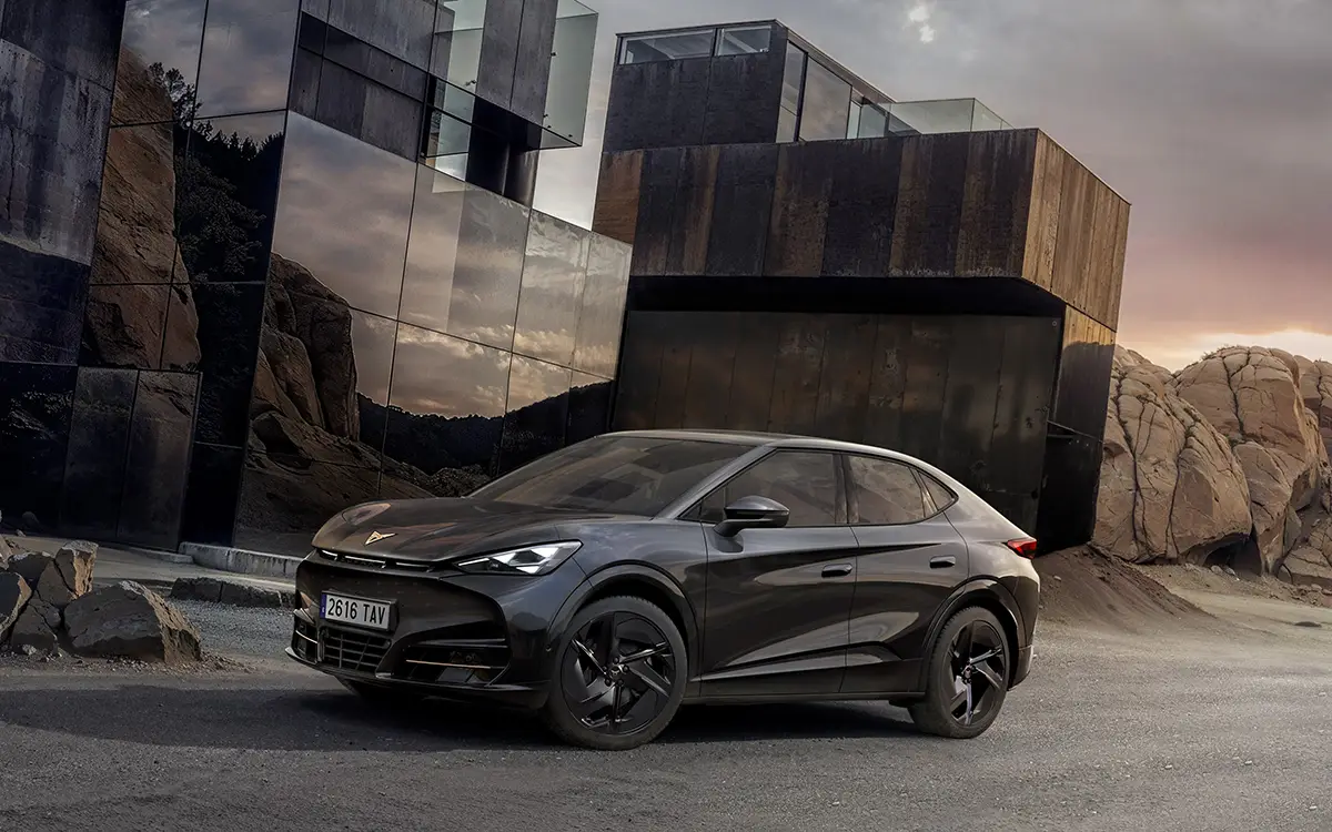 Der rein elektrische Cupra Tavascan Black Edition in der tiefschwarzen Lackierung Midnight Black und mit schwarzen 21-Zoll-Alufelgen parkt auf einer asphaltierten Strasse vor einer modernen, kantigen Gebäudearchitektur aus dunklem Metall.