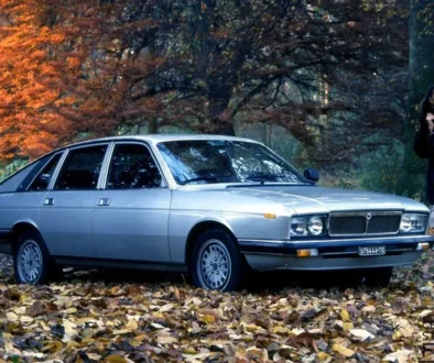Klassische Lancia Gamma Schrägheck-Limousine aus dem Jahr 1976 im Farbton Silbermetallic, geparkt auf herbstlichem Laub neben einer elegant gekleideten Frau.