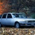 Klassische Lancia Gamma Schrägheck-Limousine aus dem Jahr 1976 im Farbton Silbermetallic, geparkt auf herbstlichem Laub neben einer elegant gekleideten Frau.