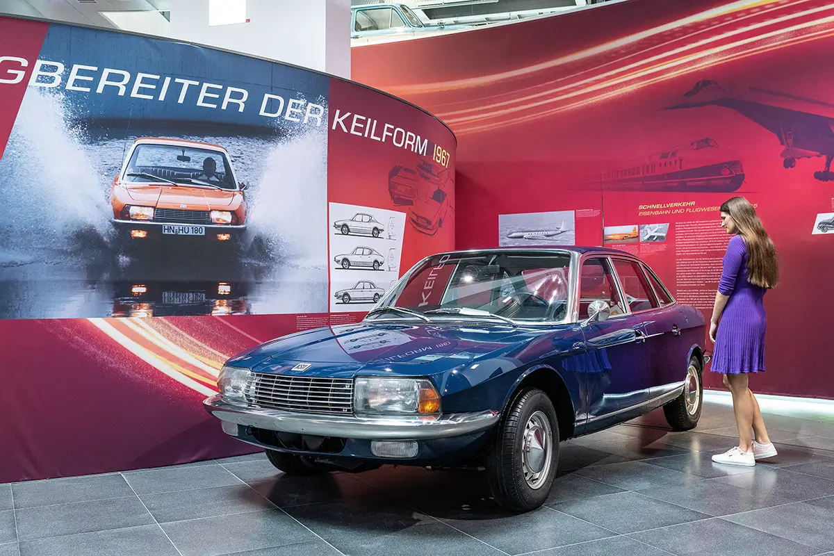 Eine dunkelblaue Limousine des Typs NSU Ro 80 aus dem Jahr 1967 ausgestellt im Werk vor einer roten Messewand mit der großen Aufschrift „Wegbereiter der Keilform“.