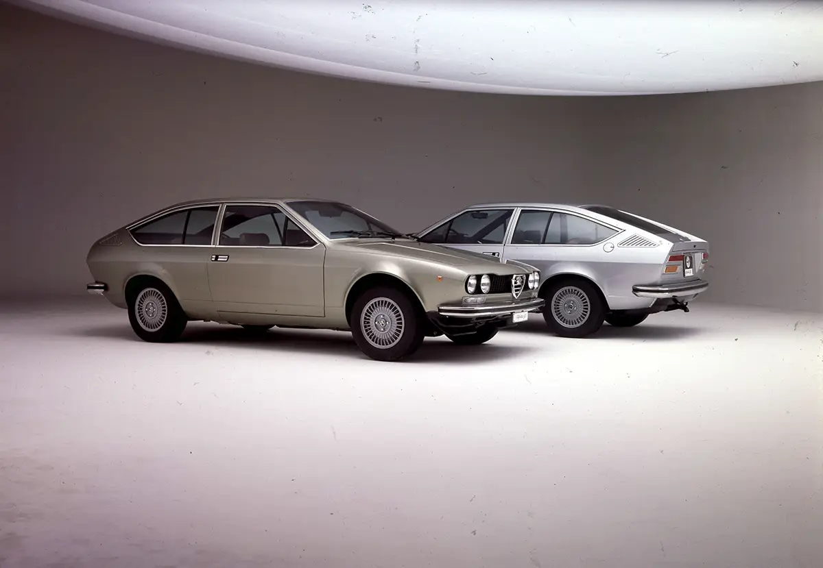 Zwei klassische, silberfarbene Alfa Romeo Alfetta GT Sportcoupés der 1970er Jahre im direkten Profilvergleich vor weißem Hintergrund im Fotostudio.