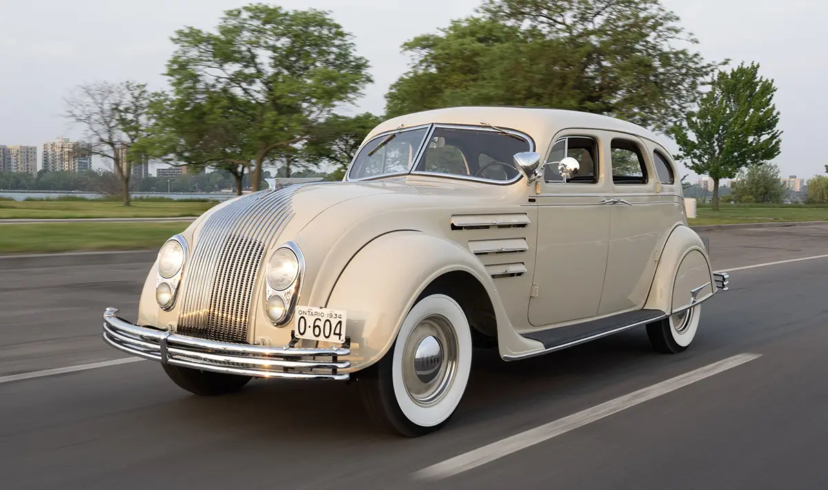 Ein historischer, cremefarbener Chrysler Airflow Sedan aus dem Jahr 1934 bei einer Demonstrationsfahrt auf einer asphaltierten Straße zur Veranschaulichung des frühen Stromliniendesigns.