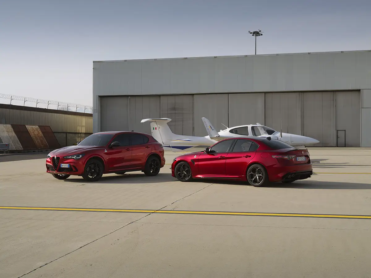 Ein rotes Alfa Romeo Stelvio SUV und eine rote Giulia Limousine in den sportlichen High-Performance-Versionen Quadrifoglio nebeneinander auf einem Flugplatzgelände vor einem Propellerflugzeug.