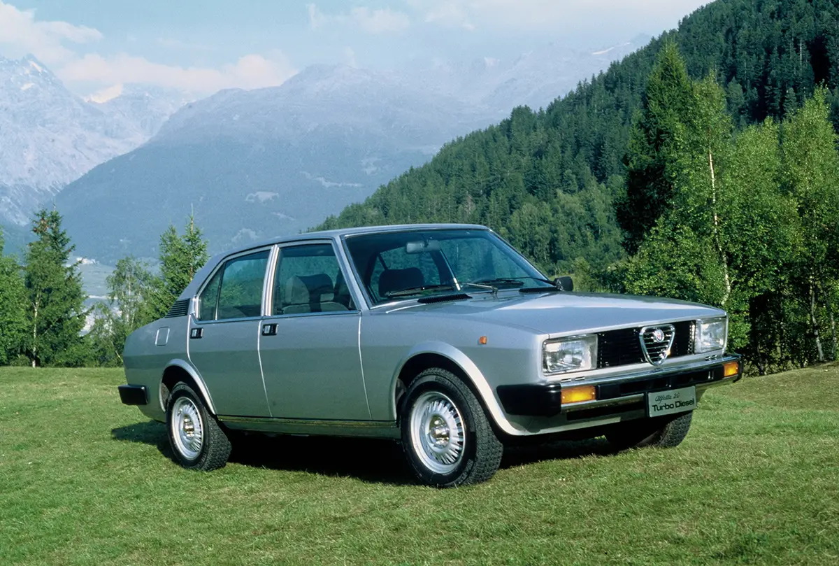 Klassische viertürige Alfa Romeo Alfetta Limousine in Silbermetallic auf einer grünen Rasenfläche vor einer alpinen Bergkulisse.