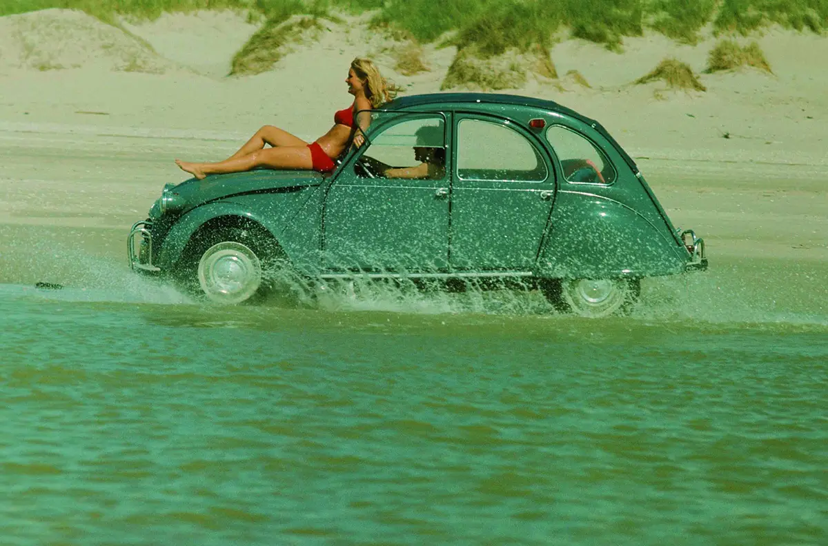 Eine smaragdgrüne Citroën 2 CV „Ente“ bei einer Fahrt durch flaches Meerwasser an einem Sandstrand, während eine Person im roten Bikini auf der Motorhaube liegt.