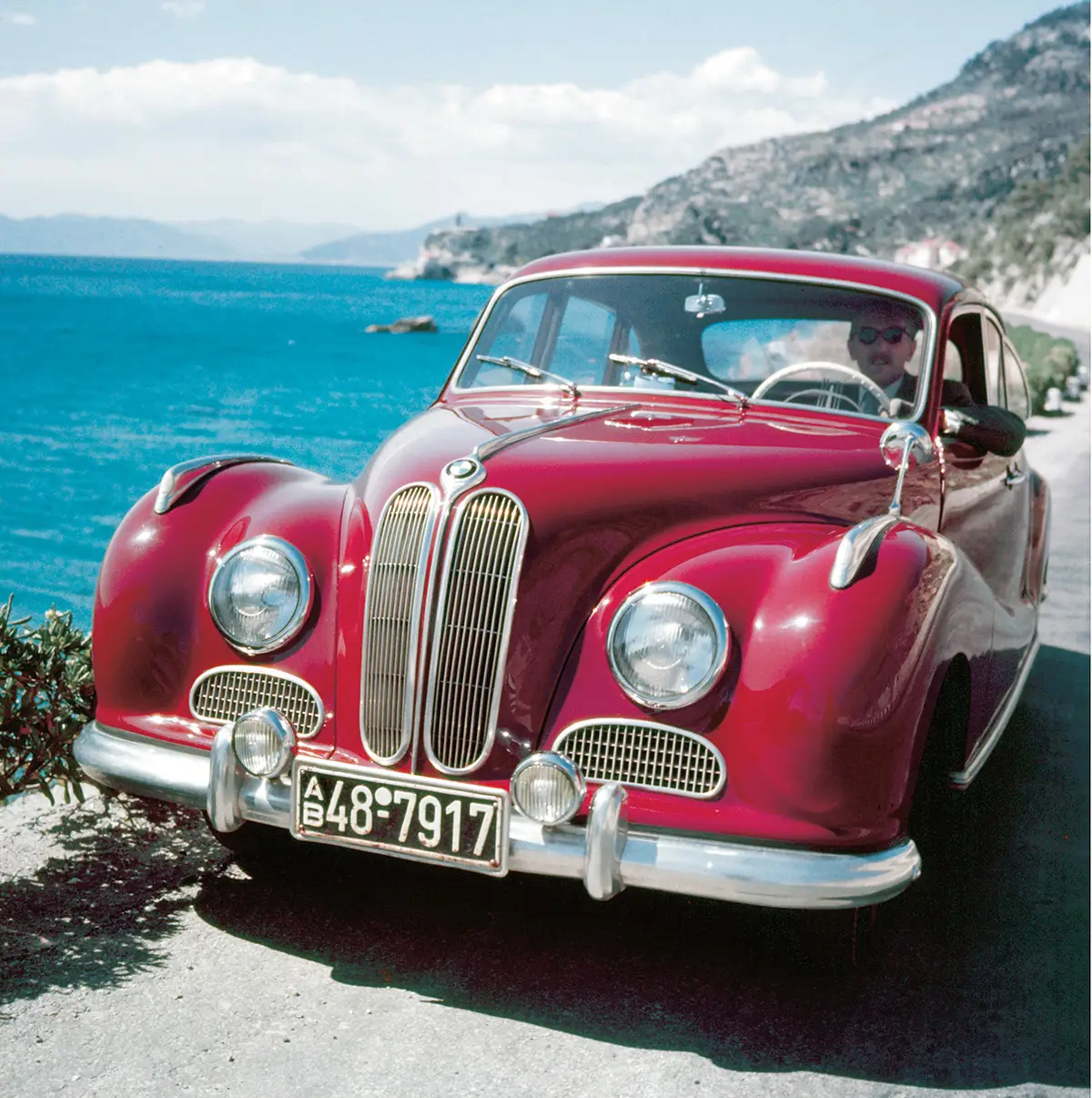 Frontalansicht eines glänzenden, weinroten BMW 501 „Barockengel“ (1951) mit opulent geschwungenen Kotflügeln und der charakteristischen Doppelniere an einer sonnigen Küstenstraße.