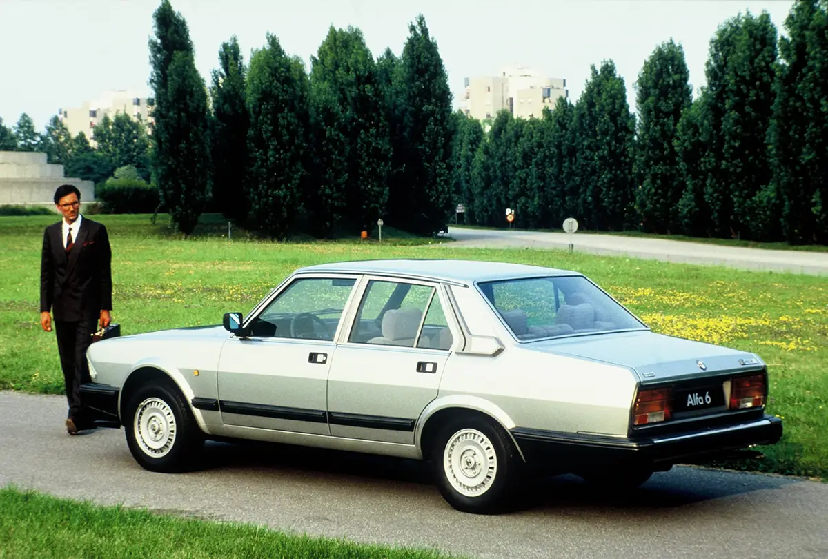 Die Heckseitenansicht einer silbernen Alfa 6 Luxuslimousine aus dem Jahr 1979, neben der ein Geschäftsmann im Anzug steht.