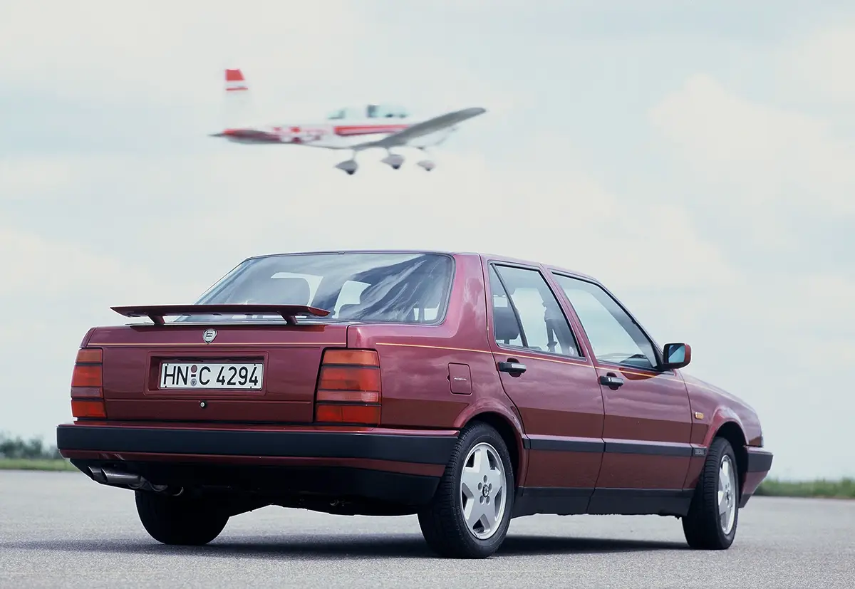 Heckansicht des weinroten Lancia Thema 8.32 mit ausfahrbarem Heckspoiler auf einer Startbahn unter einem tief fliegenden Flugzeug.