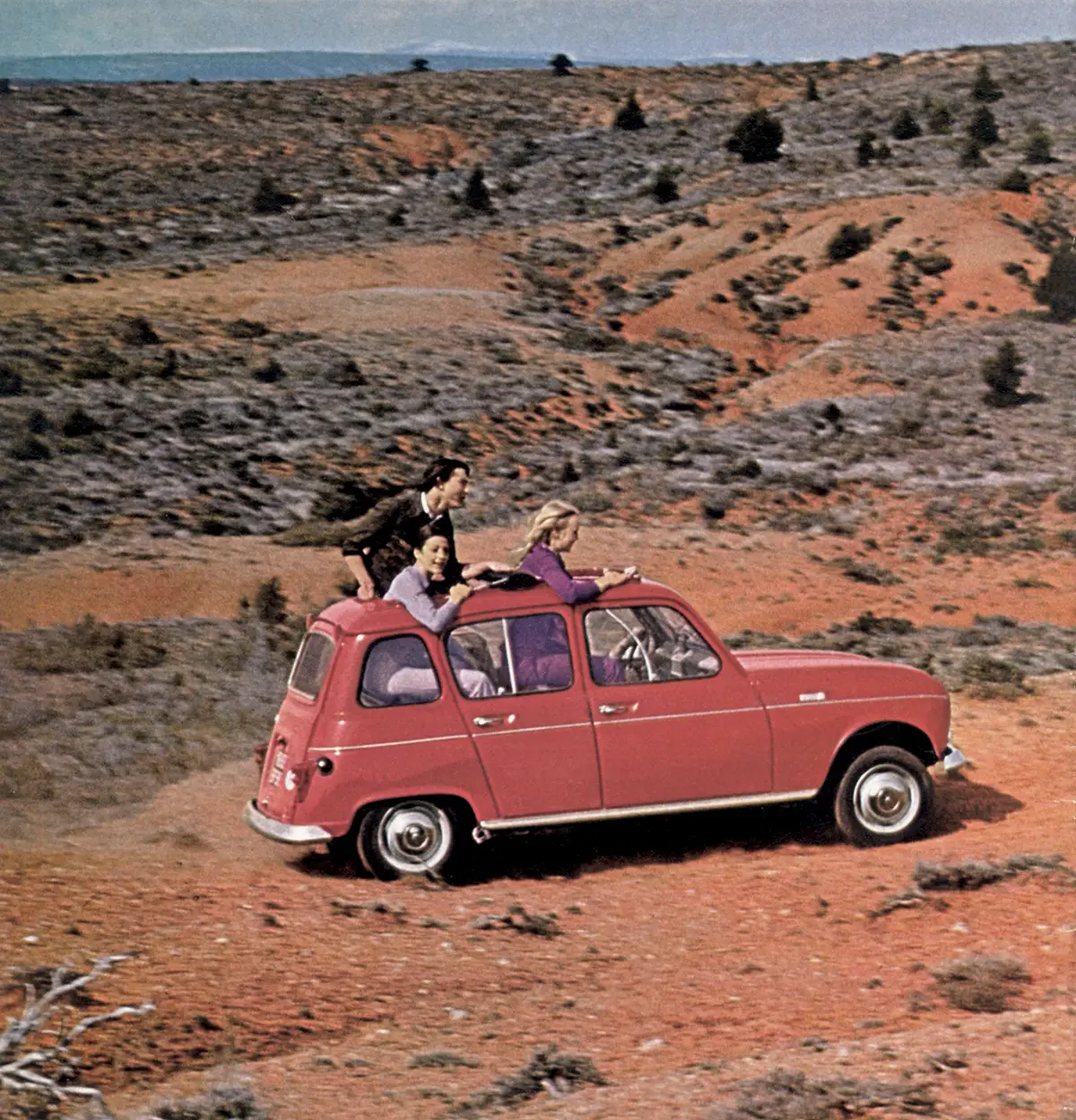 Ein rosaroter, minimalistischer Renault R4 aus dem Jahr 1961 fährt vollbesetzt über eine unbefestigte Piste im Gelände, während Fahrgäste aus dem geöffneten Faltdach herausschauen.