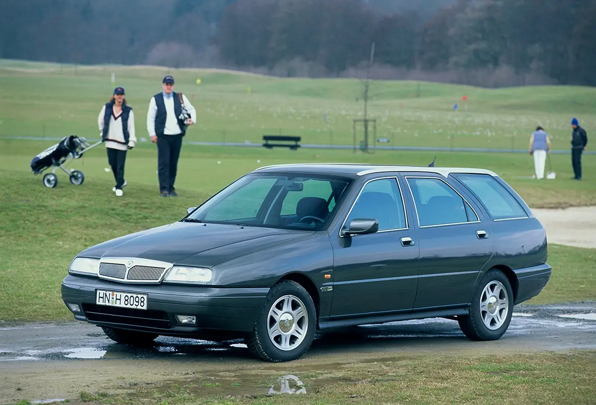 Ein dunkelgrauer Lancia Kappa Kombi (Station Wagon) auf dem Asphaltweg eines grünen Golfplatzes mit Golfern im Hintergrund.