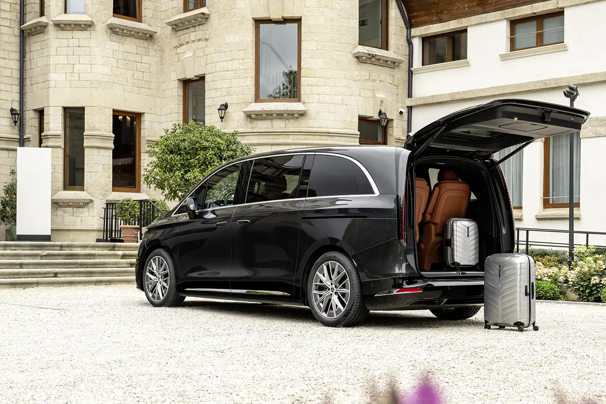 Seiten-Heck-Profil des Mercedes VLE vor einem herrschaftlichen Anwesen mit komplett elektrisch geöffneter Heckklappe, die den Blick auf den großen Laderaum und silberne Koffer freigibt