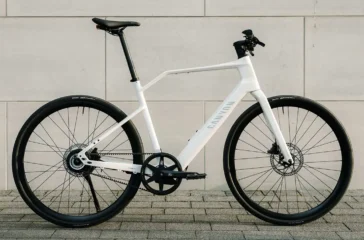 Seitenprofil des Canyon Roadlite:On CF E-Bikes mit weissem Carbonrahmen, integrierter Starrgabel, geradem Lenker und unauffälligem Bosch-Nabenmotor vor einer grauen Betonwand