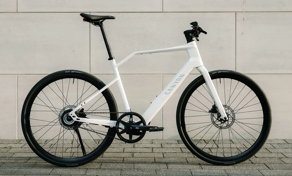 Seitenprofil des Canyon Roadlite:On CF E-Bikes mit weissem Carbonrahmen, integrierter Starrgabel, geradem Lenker und unauffälligem Bosch-Nabenmotor vor einer grauen Betonwand