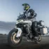 Ein Motorradfahrer mit Helm und schützender Enduro-Bekleidung fährt auf der weiß-grauen Reiseenduro CFMoto 800MT-ES samt montierten Aluminium-Seitenkoffern über eine unbefestigte Piste an einer Gewässerkulisse.