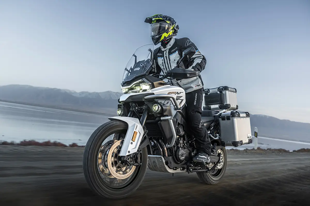 Ein Motorradfahrer mit Helm und schützender Enduro-Bekleidung fährt auf der weiß-grauen Reiseenduro CFMoto 800MT-ES samt montierten Aluminium-Seitenkoffern über eine unbefestigte Piste an einer Gewässerkulisse.