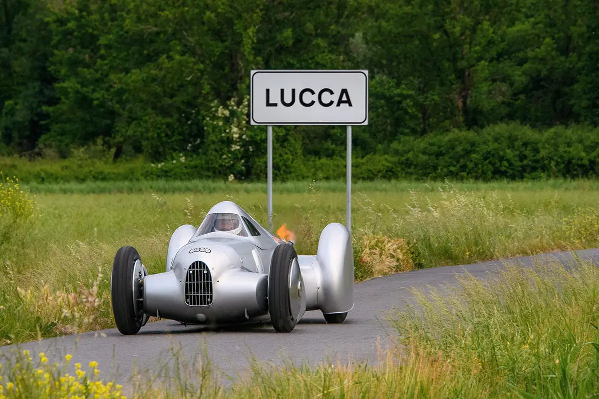 Der rekonstruierte Auto Union Typ A Stromlinien-Rekordwagen steht im Grünen unmittelbar vor einem weissen italienischen Ortsschild mit der Aufschrift „LUCCA“.